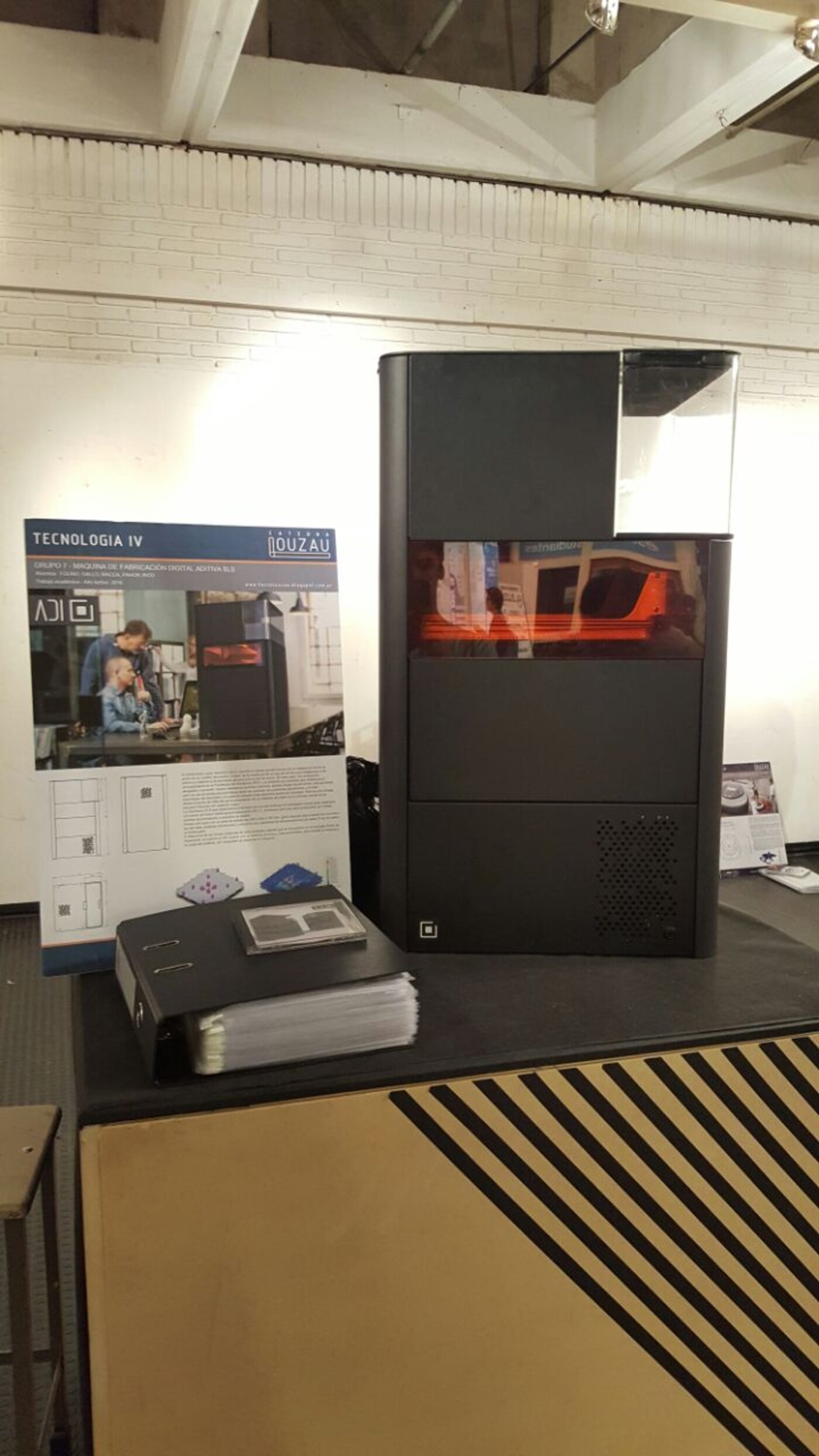 The ADI machine on a presentation table during final project review, next to a project poster bound documentation and product samples