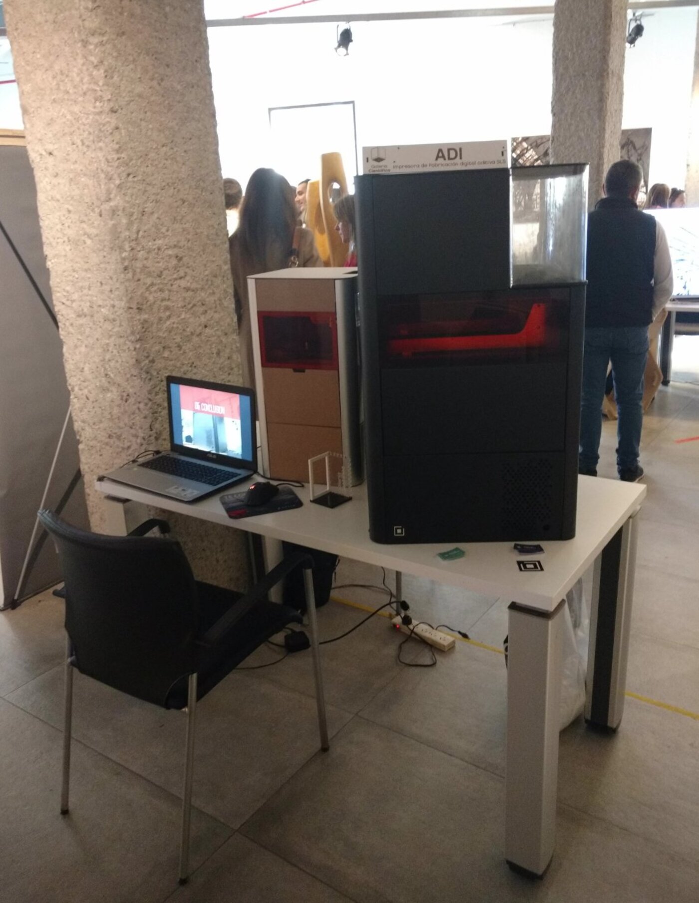 ADI machine on display at an exhibition, on a table with a laptop showing the project presentation, with visitors in the background